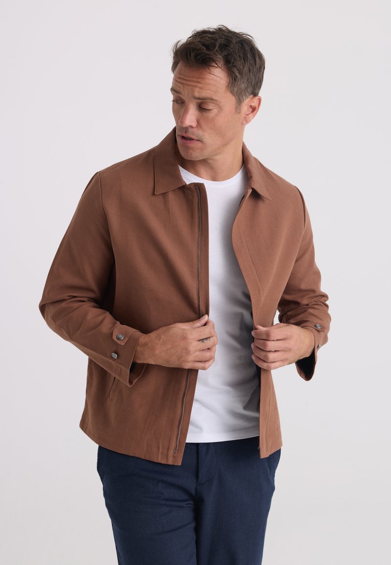 Homme ajustant une veste marron à fermeture éclair portée sur une chemise blanche et un pantalon bleu devant un fond clair uni.