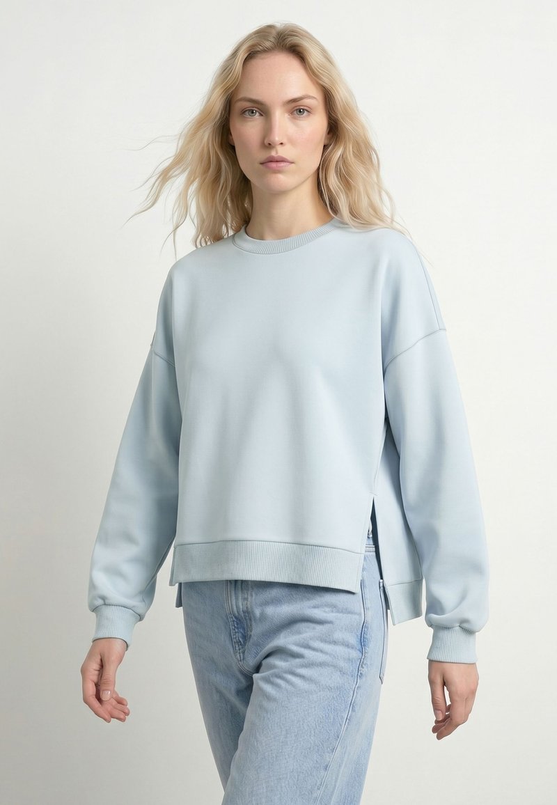 Femme blonde portant un sweat-shirt oversize bleu clair et un jean bleu clair, debout devant un fond blanc uni.