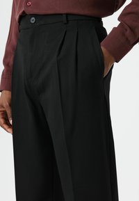 Pantalon noir plissé à coupe ajustée, doté d'une fermeture à bouton et de poches latérales. Confectionné en tissu lisse avec une finition mate.