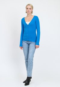 Cardigan blu brillante, con bottoni, scollo a V, maniche lunghe e polsini a costine, indossato sopra una maglietta bianca, abbinato a jeans chiari e stivali neri.
