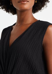 Czarny top bez rękawów z głębokim dekoltem w kształcie litery V, wykonany z fakturowanej tkaniny z pionowymi fałdami. Złote kolczyki w kształcie kropli uzupełniają stylizację.