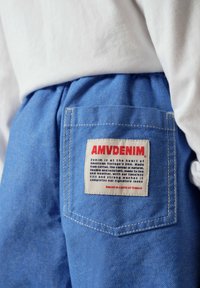 Poche de pantalon en denim bleu avec une étiquette blanche montrant un texte rouge et noir indiquant AMVDENIM et une description du produit en petits caractères.