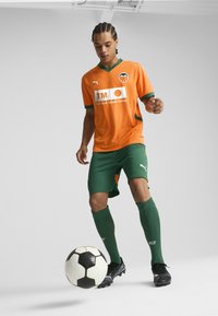 Oranžový futbalový dres so zelenými akcentmi, zelené šortky a čierne kopačky. Dres má na prednej strane logo a sponzorskú značku.
