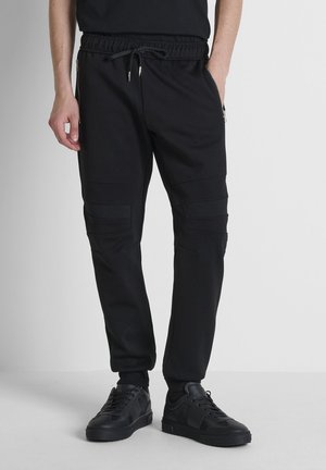 Man draagt zwarte joggerbroek met trekkoord in de taille en ritssluiting zijzakken, gecombineerd met zwarte sneakers, staand op een lichtgrijze vloer.