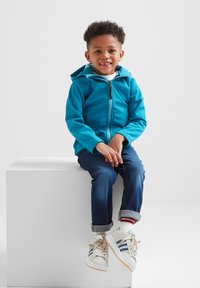 Bright blue zip-up jacket with a hood, worn over a white shirt, paired with dark blue jeans and beige sneakers with blue and red stripes.