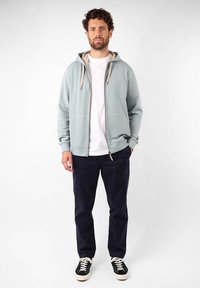 Haut de survêtement zippé bleu clair avec des cordons beiges, porté sur un t-shirt blanc, associé à un pantalon bleu marine et des baskets noires. Design et coupe décontractés.
