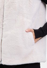 Bílá vestu z umělé kožešiny s měkkou texturou, která má boční kapsy a černý dlouhý rukáv uvnitř. Zapínání na zip v přední části.