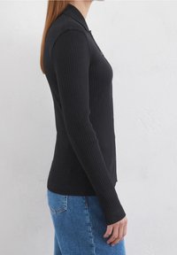 Kvinna som bär en åtsittande svart ribbad knapplångärmad cardigan och blå jeans, stående på sidan mot en enfärgad vit vägg.