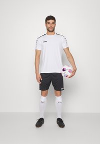 Balta sportinė apranga su juodais akcentais ir logotipu, derinama su juodomis šortais, baltais kelių ilgio kojinėmis ir juodais sportbačiais, laikantis futbolo kamuolį.