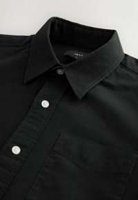 Must musta shirt, mis on valmistatud puuvillast. Omab teravat kraed, valgeid nuppe ja rinna taskut. Sile tekstuur klassikalise disainiga.
