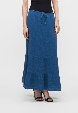 Femme portant une longue jupe bleue texturée avec une taille à cordon et un ourlet à volants, associée à des sandales noires à talons ouverts.