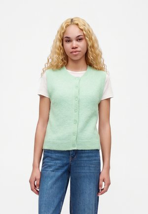 Ung kvinde med krøllet blondt hår bærer en lysegrøn knapvest over en hvid t-shirt og blå jeans, stående mod en ensfarvet baggrund.