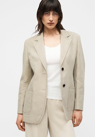 Γυναίκα που φοράει μπεζ σακάκι με δύο κουμπιά πάνω από άσπρο τοπ και ταιριαστό μπεζ παντελόνι, στέκεται μπροστά σε ένα απλό ανοιχτό φόντο.