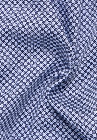 Tessuto a trama con un motivo geometrico di piccoli cerchi blu contornati di navy su uno sfondo azzurro, con una consistenza liscia.