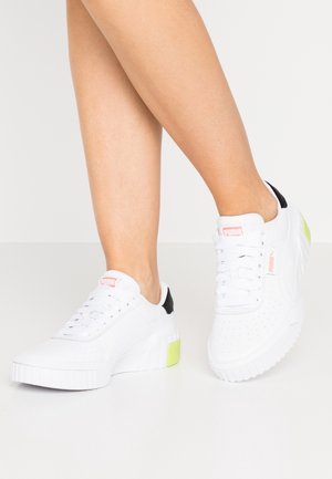 Ben iført hvide Puma-sneakers med sorte hæl-accenter og lysegrønne detaljer på tykke såler mod en ensfarvet hvid baggrund.