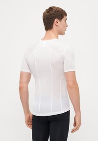 T-shirt de sport ajusté blanc à manches courtes, en tissu texturé, avec des lignes de couture diagonales contrastantes. Porté avec des leggings de sport noirs.