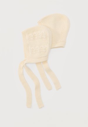 Dve krémovej farby pletené detské čiapky, jedna s kvetinovými vzormi v zapletenom vzore a dlhými stuhami, položené na bielom pozadí.