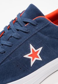 Zapatillas de ante azul marino con detalles y cordones rojos, que presentan un logo de estrella blanca. Parte superior texturizada con perforaciones y suela de goma blanca.
