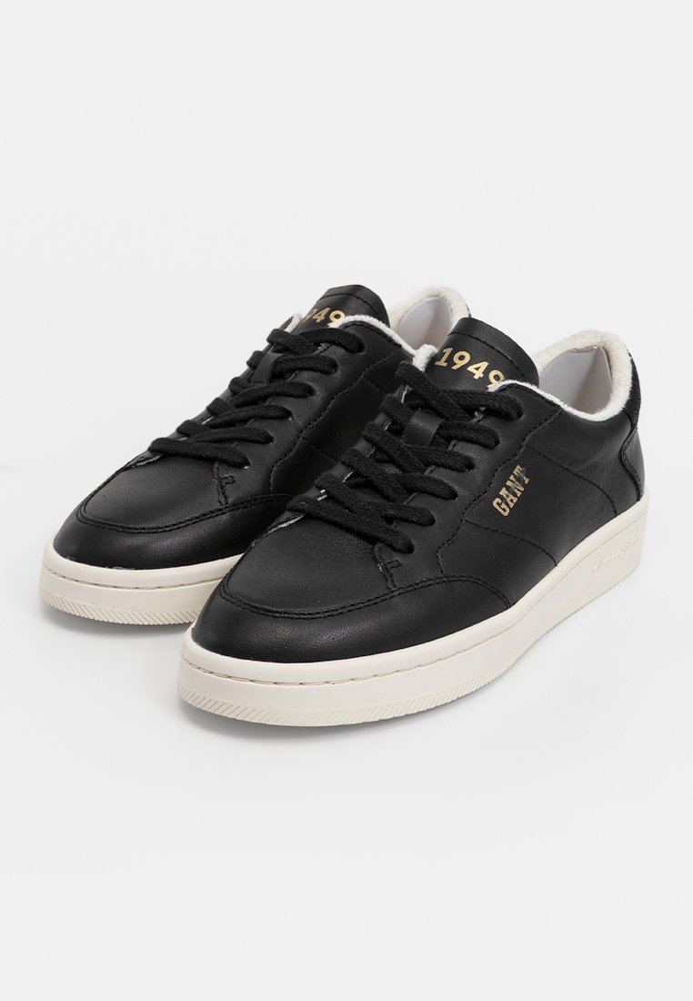 Schwarze Leder-Sneaker mit weißen Gummisohlen, ausgestattet mit schwarzen Schnürsenkeln und einer weichen weißen Innenausstattung. Goldene Akzente umfassen den Markennamen und das Jahr "1949".