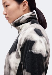Άτομο με σκούρα μαλλιά δεμένα πίσω φοράει fleece μπουφάν με μοτίβο σε μαύρο, άσπρο και γκρι με το λογότυπο της Columbia στο πάνω μέρος του μανικιού.