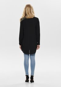 Czarna koszula z długim rękawem o prostym kroju, zestawiona z jasnoniebieskimi dżinsami. Góra ma zaokrąglony dół i zapinane mankiety.