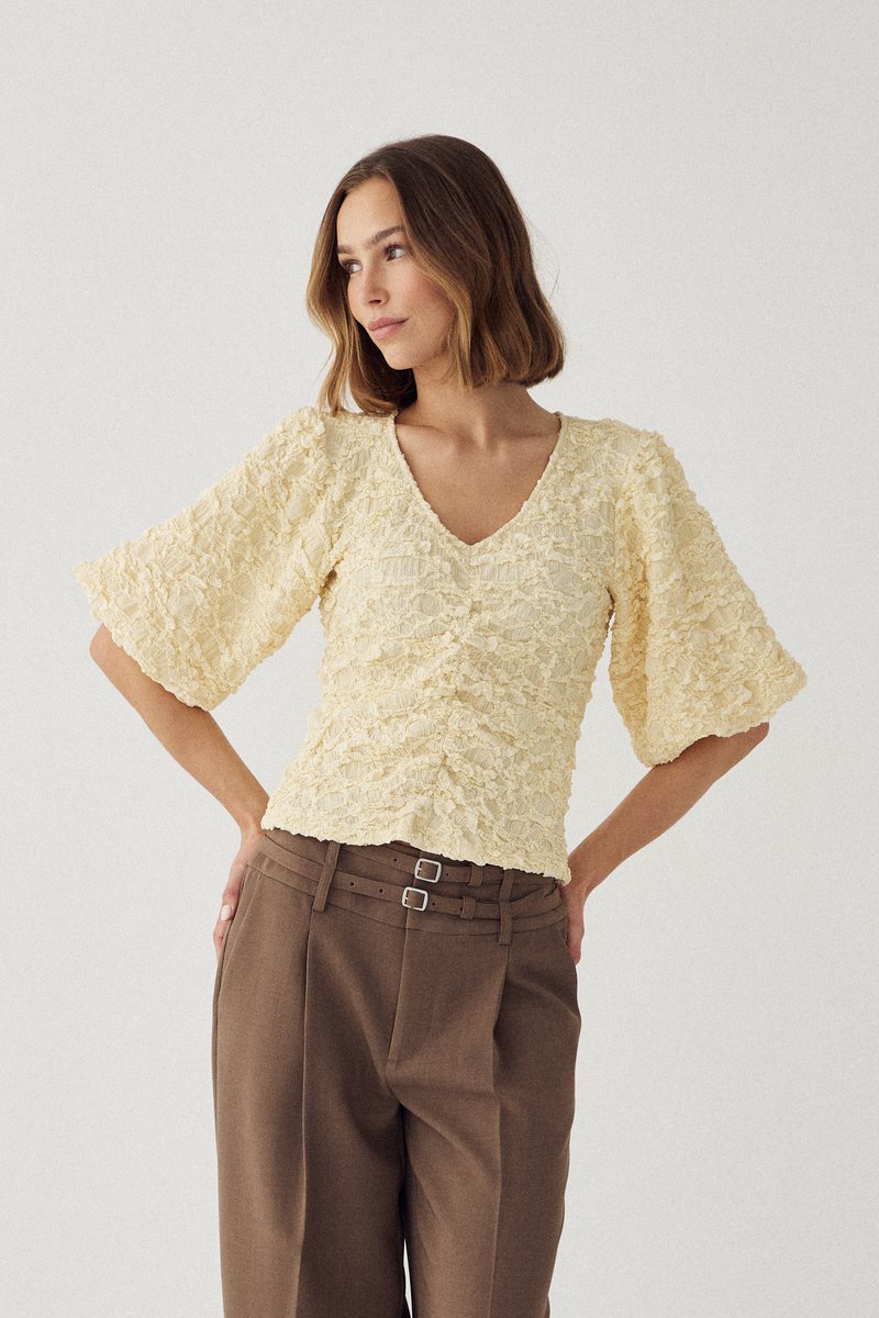 Femme portant un chemisier crème texturé à col en V avec des manches larges et un pantalon marron taille haute avec deux ceintures, posant les mains sur les hanches.