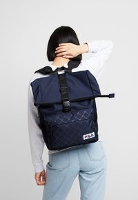 Námořnický modrý batoh s texturovaným vzorem, nastavitelnými popruhy, přední kapsou a logem FILA. Vyroben z odolného materiálu se zaklapávacím uzávěrem.