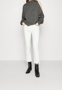 Šedý svetr se spadlými rameny a žebrovanými manžetami, doplněný bílými zvonovými džíny s neupraveným lemem. Černé kotníkové boty dokončují outfit.