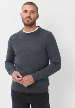 Mann mit kurzen Haaren und Bart, der einen dunkelgrauen Pullover über einem weißen Hemd trägt, vor einem einfarbigen hellen Hintergrund stehend.