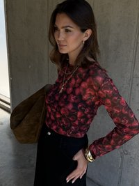 Langärmliges Oberteil mit einem roten Obstmuster, kombiniert mit einem schwarzen Rock. Die Accessoires umfassen ein rundes Goldarmband und eine braune Wildledertasche.