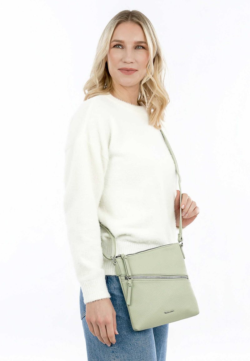 Borsa a tracolla in pelle verde chiaro con texture liscia, dotata di chiusura a zip e tracolla regolabile, indossata su un maglione bianco.