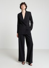 Schwarzer maßgeschneiderter Anzug mit weißen Polka-Dots, bestehend aus einem Blazer mit tiefem V-Ausschnitt und weit geschnittenen Hosen. Glatte Textur, minimalistisches Design. High-Heels.