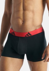 Μαύρα μποξεράκια με κόκκινη ελαστική μέση που διαθέτει μαύρες λωρίδες με λογότυπο. Το υλικό φαίνεται λείο με εφαρμοστό σχέδιο και ελάχιστες ραφές.