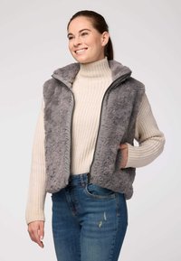 Pelēks mākslīgā kažokāda vests ar augstu apkakli, ar rāvējsl�ēdzēju un sānu kabatām, uzvilkts virs bēša adīta turtlenecka un ziliem džinsiem. Mīksta tekstūra.