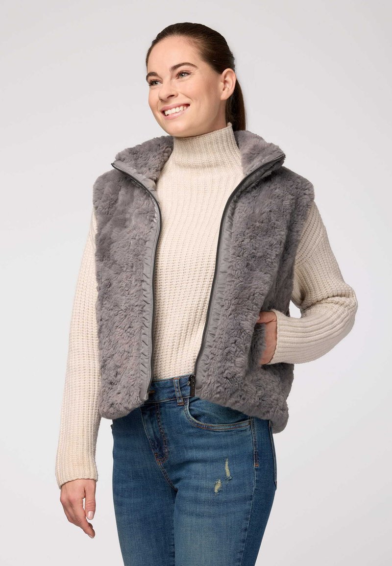 Pelēks mākslīgā kažokāda vests ar augstu apkakli, ar rāvējslēdzēju un sānu kabatām, uzvilkts virs bēša adīta turtlenecka un ziliem džinsiem. Mīksta tekstūra.