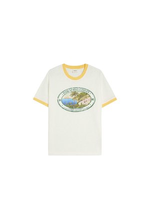 Mann mit lockigem Haar, der orange reflektierende Sonnenbrille, ein weißes T-Shirt mit Küstenmotiv und gestreifte Hose trägt und an einer bunten blau-grünen Wand lehnt.