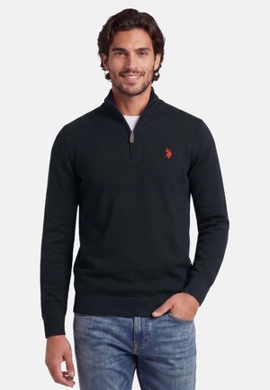 Man smiling wearing a black quarter-zip sweater with a red logo and light blue jeans, standing against a white background.