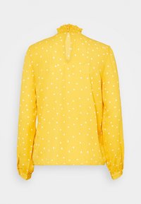 Gul blus med vita prickar, långa ärmar, volang i halsen och nyckelhål på baksidan. Tillverkad av lättviktsmaterial.