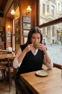 Kvinde smiler, mens hun holder en kop i en vintage-stil café med træborde, indrammede billeder på væggene og en brostensbelagt gade udenfor vinduet.