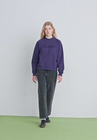 Purple sweatshirt with a crew neck and embroidered logo, paired with black wide-leg pants. Model wears black shoes and white socks.