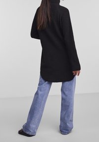 Svart ribbad polotröja med långa ärmar, i kombination med ljusblå vida jeans, sedd bakifrån.