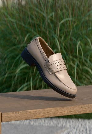 Mocassino beige in pelle con dettaglio di cinturino traforato e suola nera, sospeso sopra una panca di legno con sfondo di erba verde.