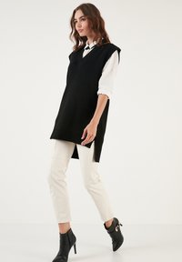 Black sleeveless knit tunic with a v-neck, layered over a white shirt, paired with cream trousers and black ankle boots.