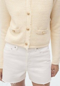 Cárdigan de punto texturizado color crema, con cierres de botones dorados y bolsillos frontales, combinado con pantalones cortos de mezclilla blancos de tiro alto y bordes deshilachados.