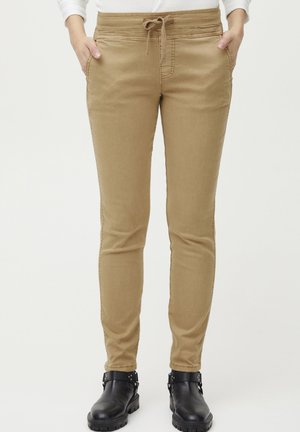 Slim-fit, tan pants with a drawstring waistband, front pockets, and subtle stitching. Worn with black ankle boots featuring metallic accents.