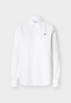 Chemise blanche à manches longues avec col et boutons, ornée d'un petit logo crocodile vert sur la poitrine gauche.