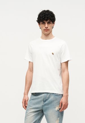 Jeune homme aux cheveux bouclés portant un t-shirt blanc uni avec un petit logo élan brodé et un jean déchiré clair.