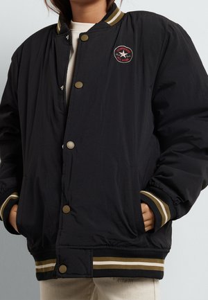 Person iført en sort varsityjakke med stribede ærmekanter og krave, med et rødt og hvidt Converse All Star logo-patch.