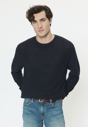 Jeune homme aux cheveux bouclés bruns portant un pull texturé foncé et un jean bleu, debout avec les mains derrière le dos sur un fond blanc.