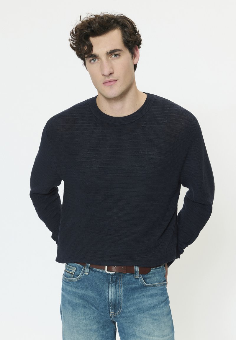 Jeune homme aux cheveux bouclés bruns portant un pull texturé foncé et un jean bleu, debout avec les mains derrière le dos sur un fond blanc.